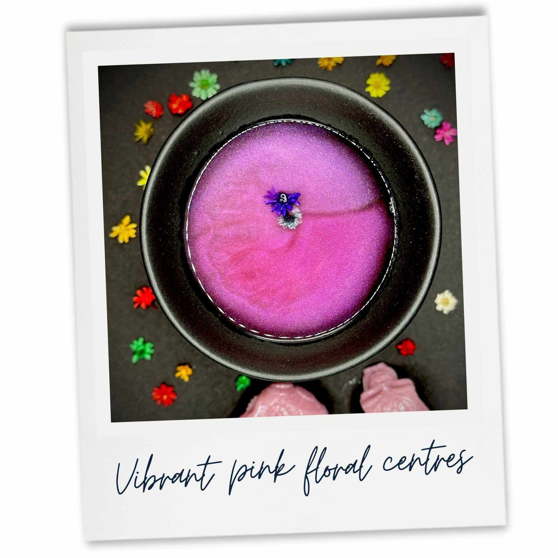 Close-up of a black wax burner containing a melted pink wax melt with a floral center. Surrounded by colourful flowers.
