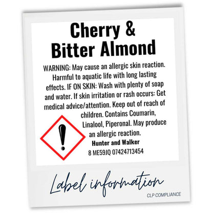 A square product label card with a white background and black text reading 'Cherry & Bitter Almond,' plus warning information, a red hazard symbol, and 'Label information' at the bottom. The card is photographed against a light gray background.