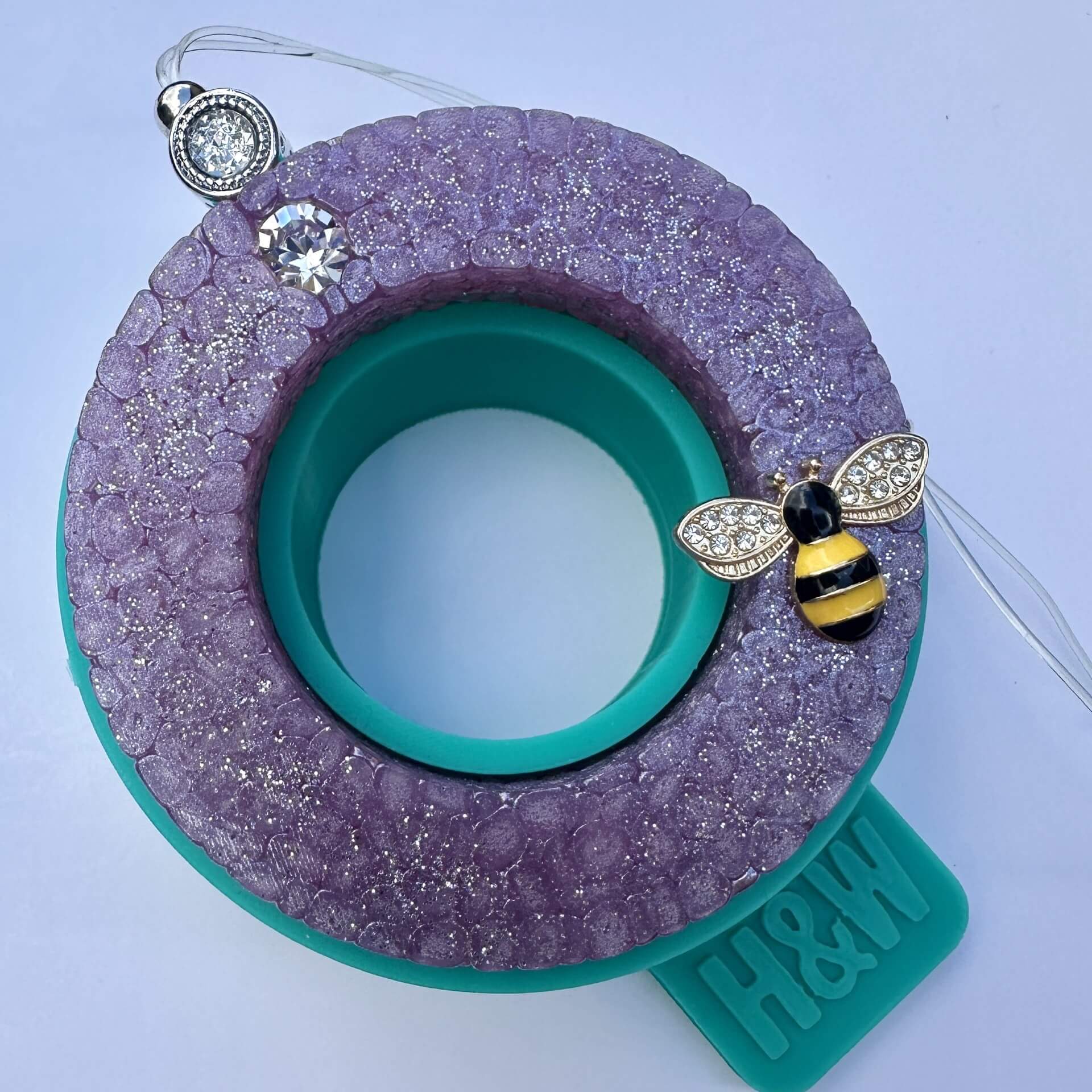 A circular purple glittery car freshener ring sat on top of the silicone mould it was created in. Decorated with a small bee charm and crystal-like accents. The wordmark 'H&W' is visible on the mould.