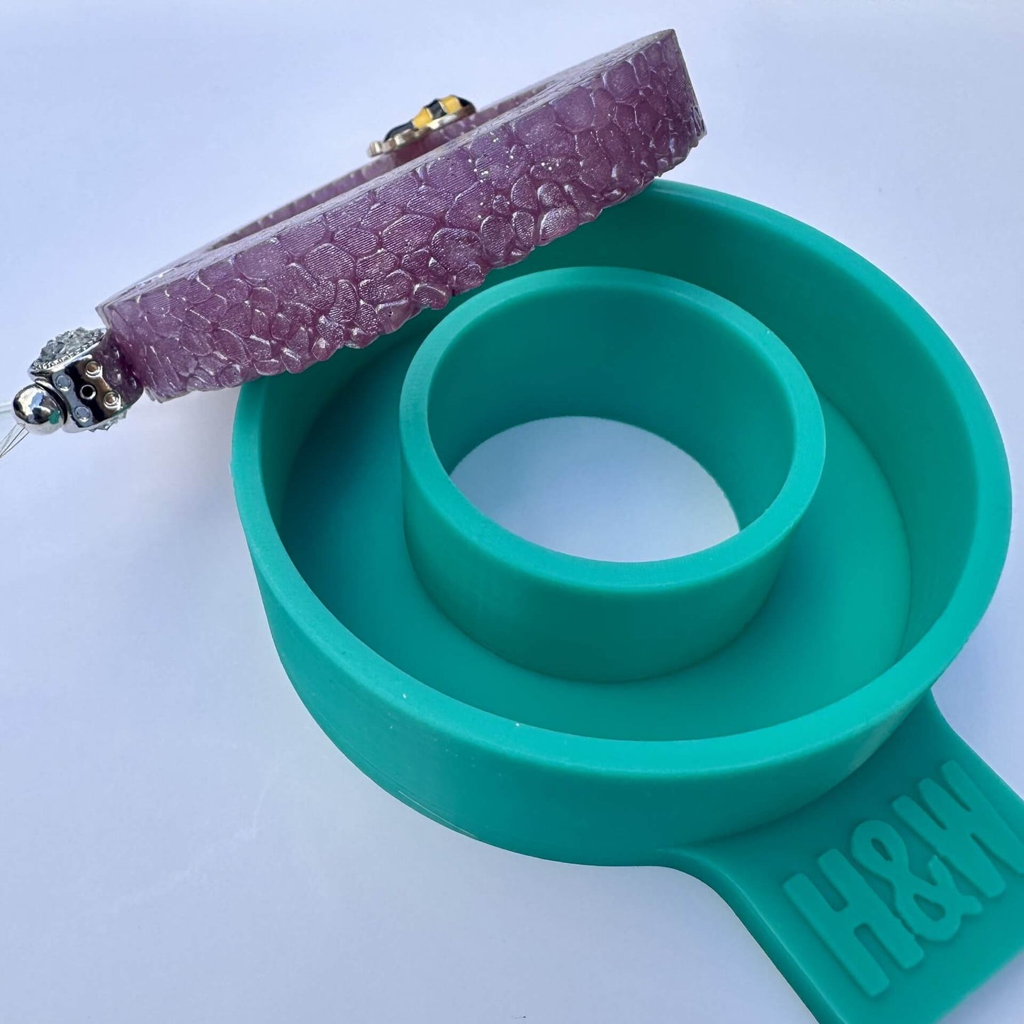 A side view of an EVA car freshie disc and it's teal green silicone ring shaped mold shown on a plain light background.